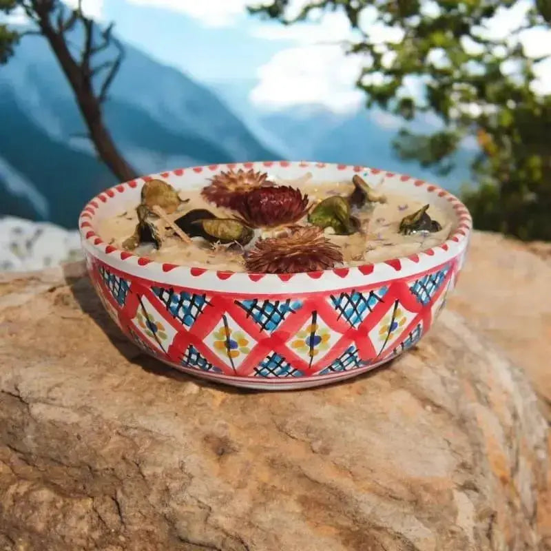 Bougie artisanale Lulu Baladine dans un bol rouge décoré de motifs géométriques, ornée de fleurs séchées, posée sur un rocher face aux montagnes.